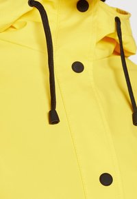 Veste imperméable jaune à finition texturée, avec boutons-pression noirs et cordons de serrage noirs. Dotée d'un col montant et d'un design sans coutures.