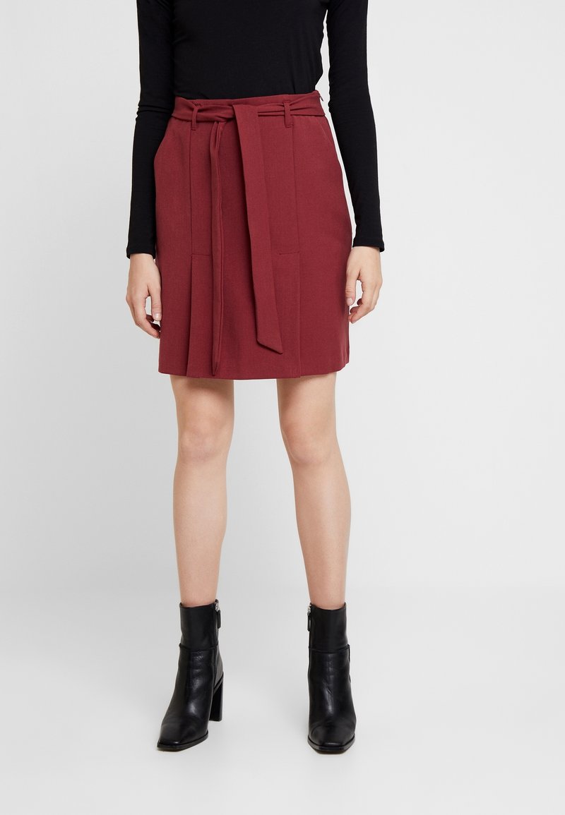 Femme portant une jupe bordeaux longueur genou avec une ceinture en tissu, un haut noir à manches longues et des bottines noires à talons épais.