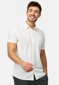 Camisa blanca de lino de manga corta con cuello abotonado, un bolsillo en la parte frontal y un acabado texturizado sutil. El modelo se está inclinando ligeramente hacia adelante.