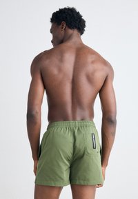 Grønne shorts laget av lett materiale med elastisk midje, sidelommer og en merket lapp bak. Synlig teksturdetalj.