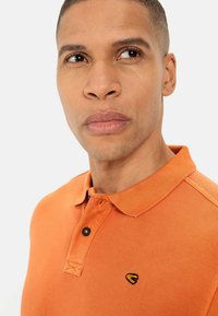 Oranje polo shirt van een gestructureerde stof, met een drieknoopsluiting en zwarte accenten. Heeft een klein geborduurd logo op de borst.