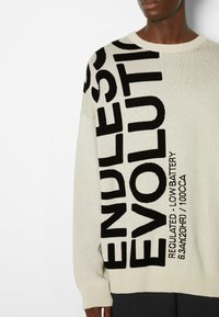 Pull crème avec texte noir indiquant "ENDLESS EVOLUTION" et instructions d'entretien. Texture tricotée, col rond, coupe décontractée.