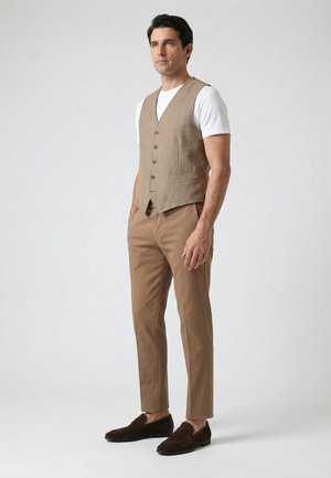 Mand iført beige vest, hvid kortærmet skjorte, beige bukser og mørkebrune loafers, står mod en ensfarvet lys baggrund.