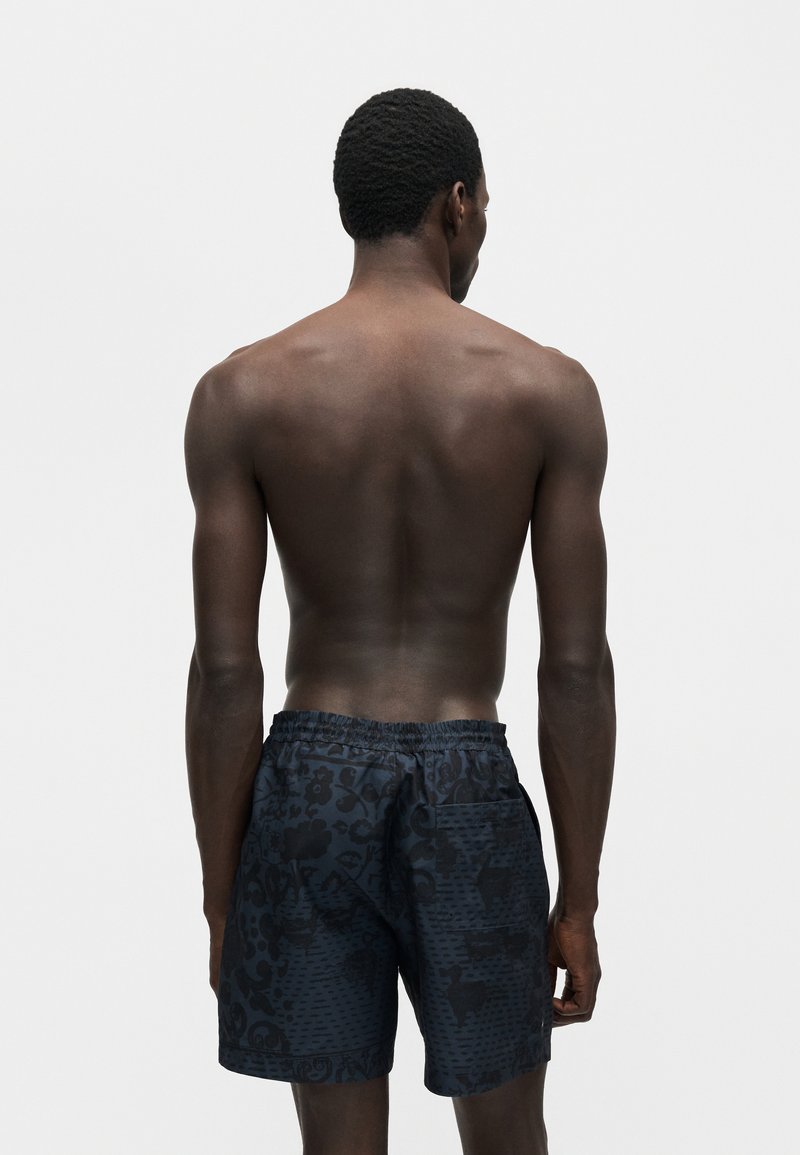 Mann mit dunkler Haut und kurzen lockigen Haaren steht oberkörperfrei, trägt dunkle gemusterte Shorts und steht mit dem Rücken zu einem einfarbigen grauen Hintergrund.