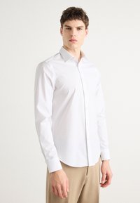 Hvit skjorte med knapper og et subtilt mønster, med klassisk krage og lange ermer, kombinert med beige bukser. Slippende tekstur.