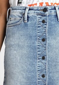 Světle modrá džínová sukně s předním zapínáním na knoflíky, dvěma bočními kapsami a dekorativním prošíváním ve zigzagovém vzoru podél zapínání na knoflíky.