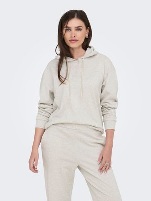 Donna con lunghi capelli castani che indossa una felpa con cappuccio beige chiaro e pantaloni abbinati, in piedi davanti a uno sfondo chiaro e uniforme.