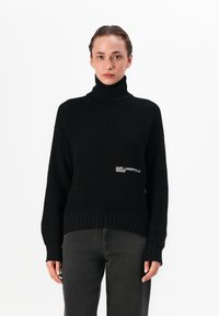 Pulover negru tricotat cu guler înalt, având o textură ribbed și un broderie cu logo pe partea stângă. Asortat cu blugi gri închis.