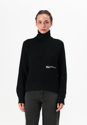 Black knitted turtleneck sweater featuring a ribbed texture and a logo embroidery on the left side. Paired with dark gray jeans.