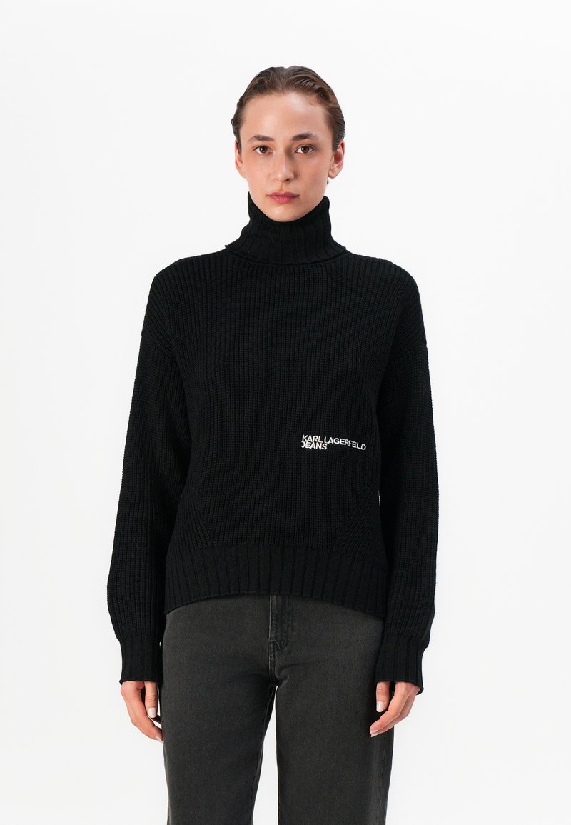 Pulover negru tricotat cu guler înalt, având o textură ribbed și un broderie cu logo pe partea stângă. Asortat cu blugi gri închis.