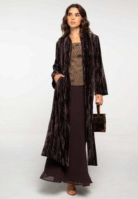 Long manteau en velours violet foncé avec des rayures verticales texturées, associé à un haut à motifs marron et une jupe marron fluide. Petit sac à main texturé.