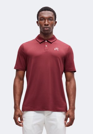 Mann trägt ein burgunderrotes Kurzarm-Polohemd mit einem kleinen weißen Logo auf der linken Brust und weiße Hose.