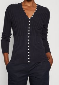 Cardigan in maglina a coste di colore blu navy con scollatura a V, chiusura con bottoni bianchi lungo la parte anteriore e rifinitura ondulata lungo il collo. Maniche lunghe.