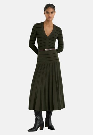 Femme portant une robe tricotée rayée vert olive avec un col en V, une ceinture marron, une jupe plissée et des bottes à talons noires, debout les mains derrière le dos.
