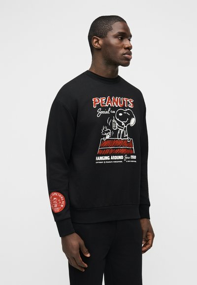 Άντρας που φοράει μαύρο φούτερ με τον Snoopy και τον Woodstock να κρατούν ποτά πάνω από το κείμενο «PEANUTS Social Club Hanging Around Since 1950».