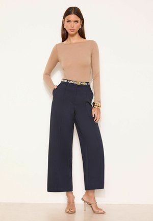 Femeie purtând un top bej cu mâneci lungi, pantaloni largi bleumarin cu o curea cu imprimeu de șarpe, brățări aurii și sandale bej cu toc, stând în picioare.