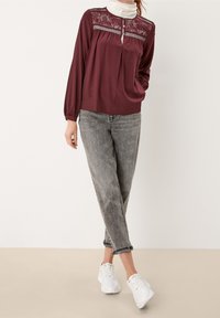 Burgunderbluse mit floralen Spitzenverzierungen am Ausschnitt, langen Ärmeln mit elastischen Bündchen, kombiniert mit grauen Jeans und weißen Sneakers.