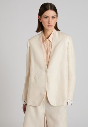 Mujer con cabello lacio y oscuro lleva una blazer beige claro, pantalones a juego y una camisa color durazno abotonada, sobre un fondo gris liso.