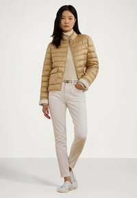 Žena oblečená v béžovej prešívanej bunde, krémovom roláku, bielych nohaviciach a bielych teniskách, stojí s jednou rukou vo vrecku.