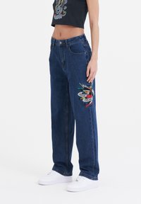 Blue denim wide-leg jeans with an embroidered bird and banner design on the left thigh. Features a high waist and front pockets.