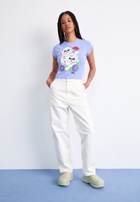 Ljuslila t-shirt med ett motiv av två katter och blommönster, kombinerat med lösa vita byxor och gröna träskor på en blå bakgrund.