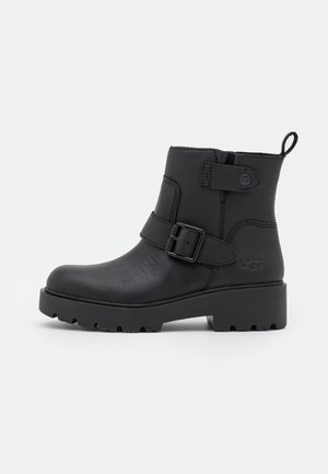 Black leather ankle boot with chunky sole, side buckle strap, ankle strap with snap button, and pull tab at the back.