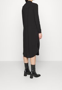 Robe chemise noire à manches longues, en tissu doux, ourlet arrondi, poignets boutonnés, détail de pli discret à l'arrière, associée à des bottines noires.