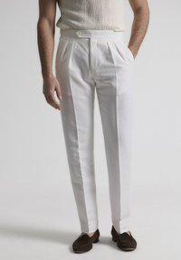 Pantalones de lino blancos con frente plisado, corte recto y cierre de un solo botón, combinados con mocasines marrón oscuro.