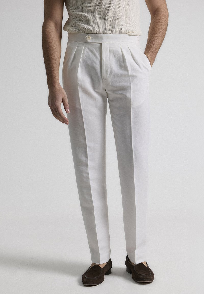 Pantalones de lino blancos con frente plisado, corte recto y cierre de un solo botón, combinados con mocasines marrón oscuro.