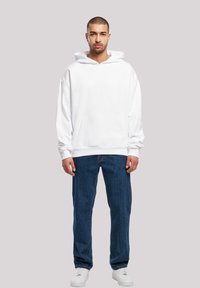 Felpa bianca oversize con tasca frontale, tessuto morbido e cappuccio con cordino, abbinata a jeans blu a gamba dritta e sneakers bianche.