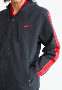 Corta-vento preto com fecho, logótipo da Nike em vermelho e riscas laterais. Tecido texturizado, capuz ajustável, bolsos frontais com fecho e design ajustado.