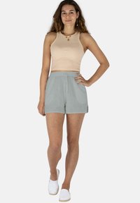 Junge Frau steht da und trägt ein beiges ärmelloses Crop-Top, hellgraue Shorts, weiße Slipper und goldenen Schmuck. Sie lächelt in die Kamera.