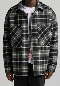 Svart och vit rutig flanellskjorta med knäppkrage, två bröstfickor och avslappnad passform, bärs över en grafisk sweatshirt.