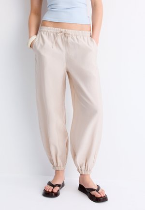 Kvinde iført beige joggebukser med elastik i taljen og opsmøgede ankler, lyseblå kort top, sorte platåsandaler og hvid armbånd.