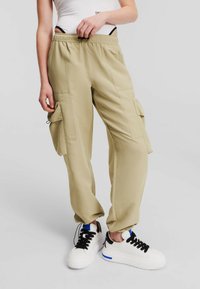 Beiga cargo-byxor med elastisk midja, sidofickor och lös passform. Bärs med svarta och vita sneakers, vilket betonar en avslappnad look.