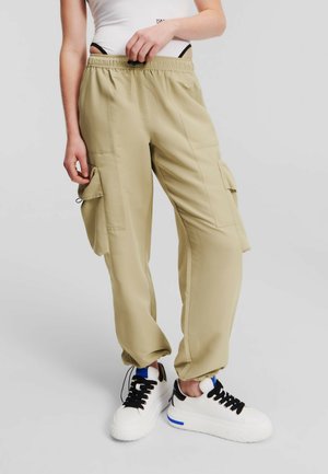 Beige cargo pants featuring an elastic waistband, side pockets, and a loose fit. Worn with black and white sneakers, emphasizing a casual look.
