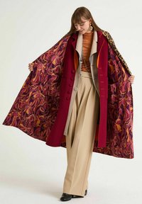 Cappotto lungo in lana color bordeaux, con fodera a motivo floreale in viola, arancione e rosa. Dettaglio di bottoni visibili e maniche larghe.
