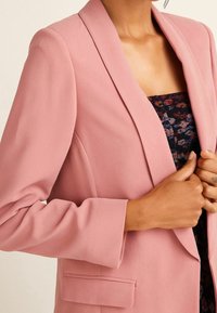 Blazer de color rosa claro con mangas largas, solapas con muesca y una tela texturizada. Presenta un bolsillo frontal y se lleva sobre una blusa de estampado.