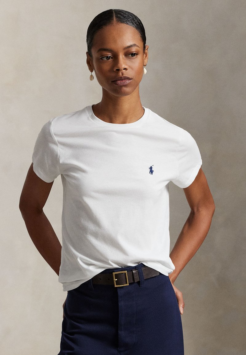 Vrouw draagt een wit t-shirt met korte mouwen met een klein donker logo op de borst en een donkere broek, staand met handen achter haar rug.