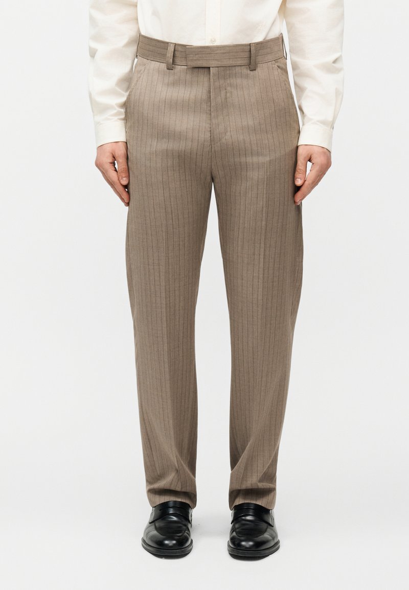 Άνδρας που φοράει μπεζ παντελόνι με ρίγες, μαύρα δερμάτινα παπούτσια και λευκό πουκάμισο με μακριά μανίκια, στέκεται με τα χέρια χαλαρά δίπλα στο σώμα.