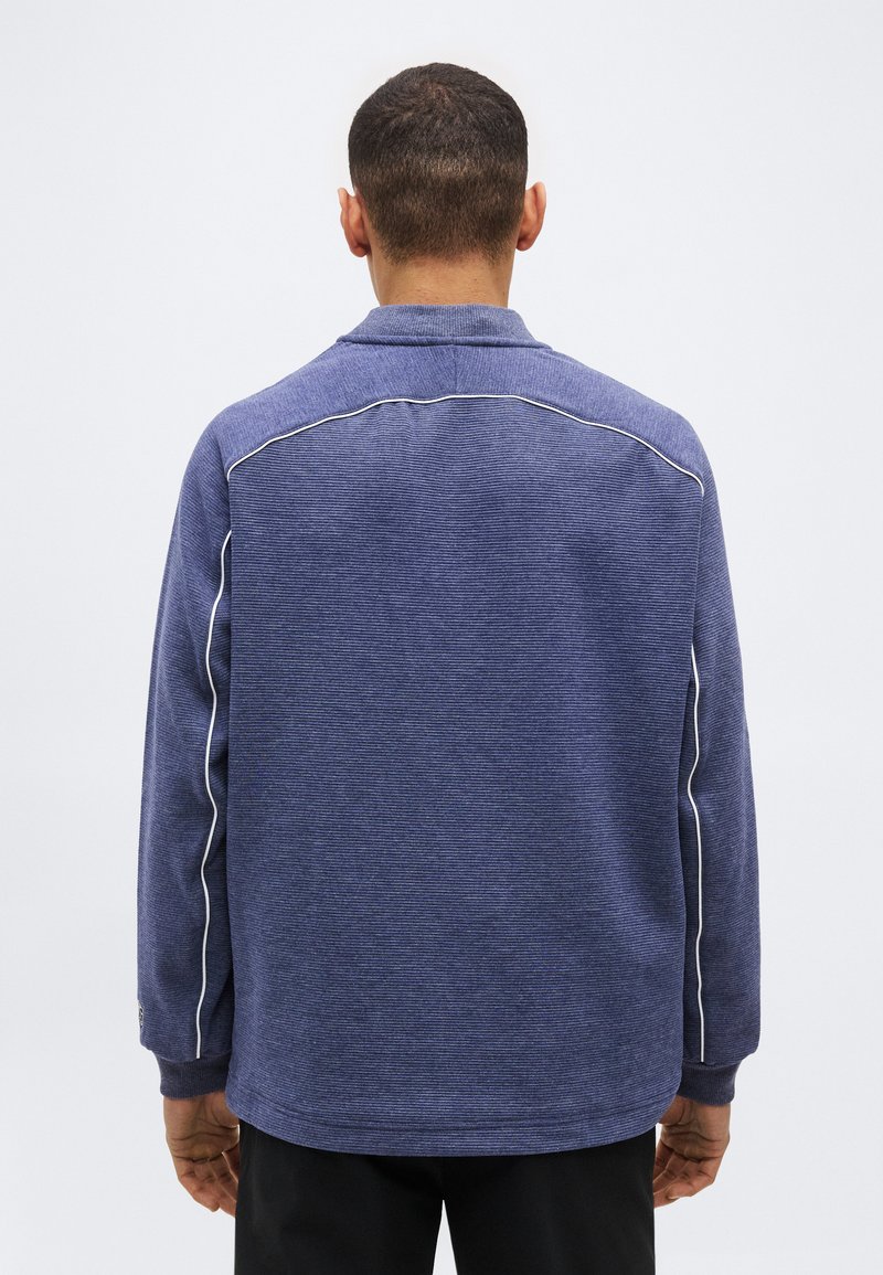 Homme de dos portant une chemise bleue à manches longues avec des passepoils blancs le long des épaules et des manches, debout devant un fond clair uni.