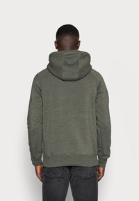 Felpa verde con un ampio cappuccio regolabile, maniche raglan e polsini e orlo a coste; tessuto morbido e strutturato; indossata con jeans scuri.