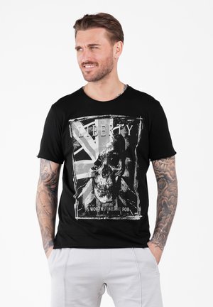 Mann mit Tattoos, der ein schwarzes T-Shirt mit einem Skull-Design und dem Text "Freiheit ist es wert, dafür zu kämpfen" trägt, steht mit Händen in den Taschen.
