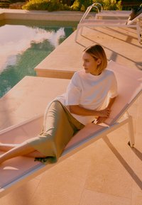 Jeune femme en haut blanc et jupe olive allongée sur une chaise longue au bord de la piscine au coucher du soleil.