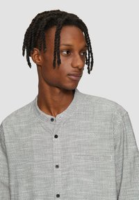 Jonge man met gevlochten haar die een lichtgrijs gestreept shirt met een bandkraag draagt, kijkt opzij tegen een effen achtergrond.