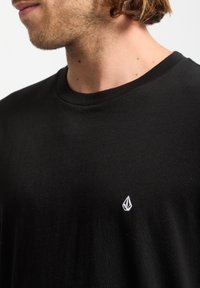 Camiseta de algodón negra con cuello redondo. Presenta un pequeño logo bordado en blanco en la parte izquierda del pecho. Textura suave.