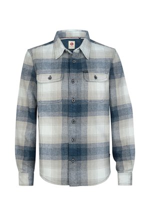 Jongen staat met één hand in zijn zak, draagt een blauw-wit geruit overhemd, blauwe spijkerbroek en witte sneakers tegen een effen achtergrond.