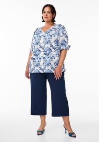 Blumige Bluse in Blau- und Weißtönen auf weißem Hintergrund, kurzärmlig, kombiniert mit marineblauen cropped Hosen und türkisfarbenen offenen Schuhen mit Absatz.