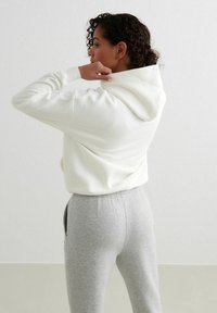 Sudadera blanca con un corte holgado y un bolsillo frontal, combinada con pantalones de chándal grises. La tela parece suave y cómoda, ideal para un uso casual.