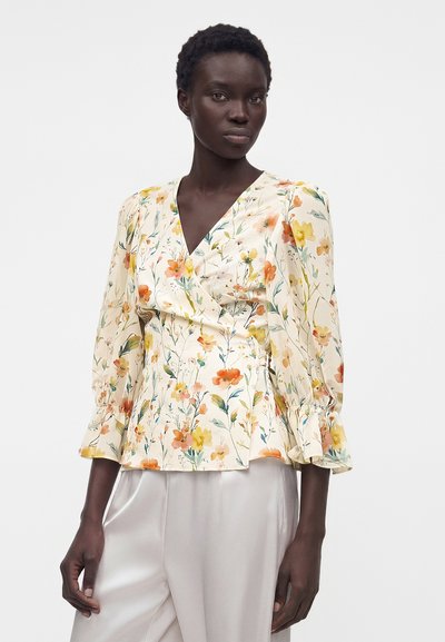 Γυναίκα με κρεμ φλοράλ μπλούζα με φουσκωτά μακριά μανίκια και βολάν στις μανσέτες, σε συνδυασμό με φαρδιά, ανοιχτόχρωμα σατινέ παντελόνια.